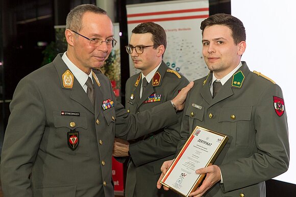 Ein Mann in Uniform hält ein Zertifikat in der Hand, ein anderer Mann in Uniform klopft ihm auf die Schulter.