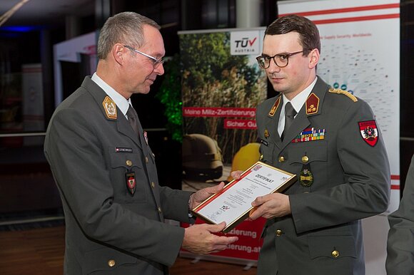 Ein Mann mit Brille in Uniform überreicht einem Mann mit Brille in Uniform ein Zertifikat.