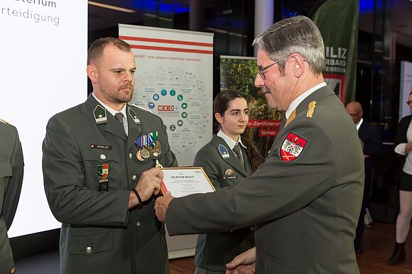 Ein Mann in Uniform und mit Brille überreicht einem Mann in Uniform ein Zertifikat.