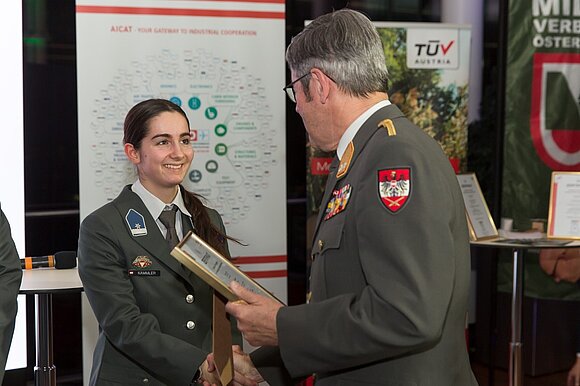 Ein Mann in Uniform überreicht einer Frau in Uniform ein Zertifikat und schüttelt ihre Hand.