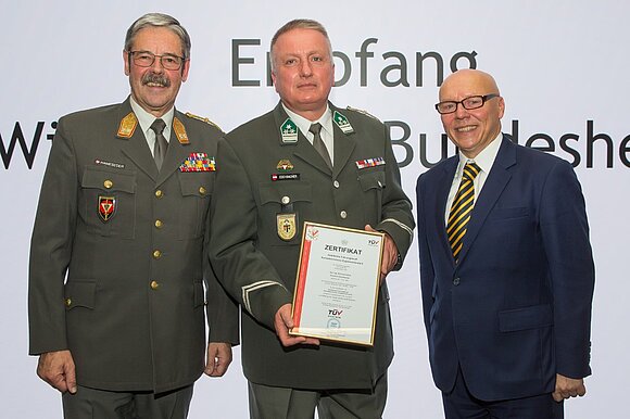 Gruppenbild mit drei Männern vor einem Plakat der WKO, zwei Männer in Uniform. Einer der Männer hält ein Zertifikat in der Hand.