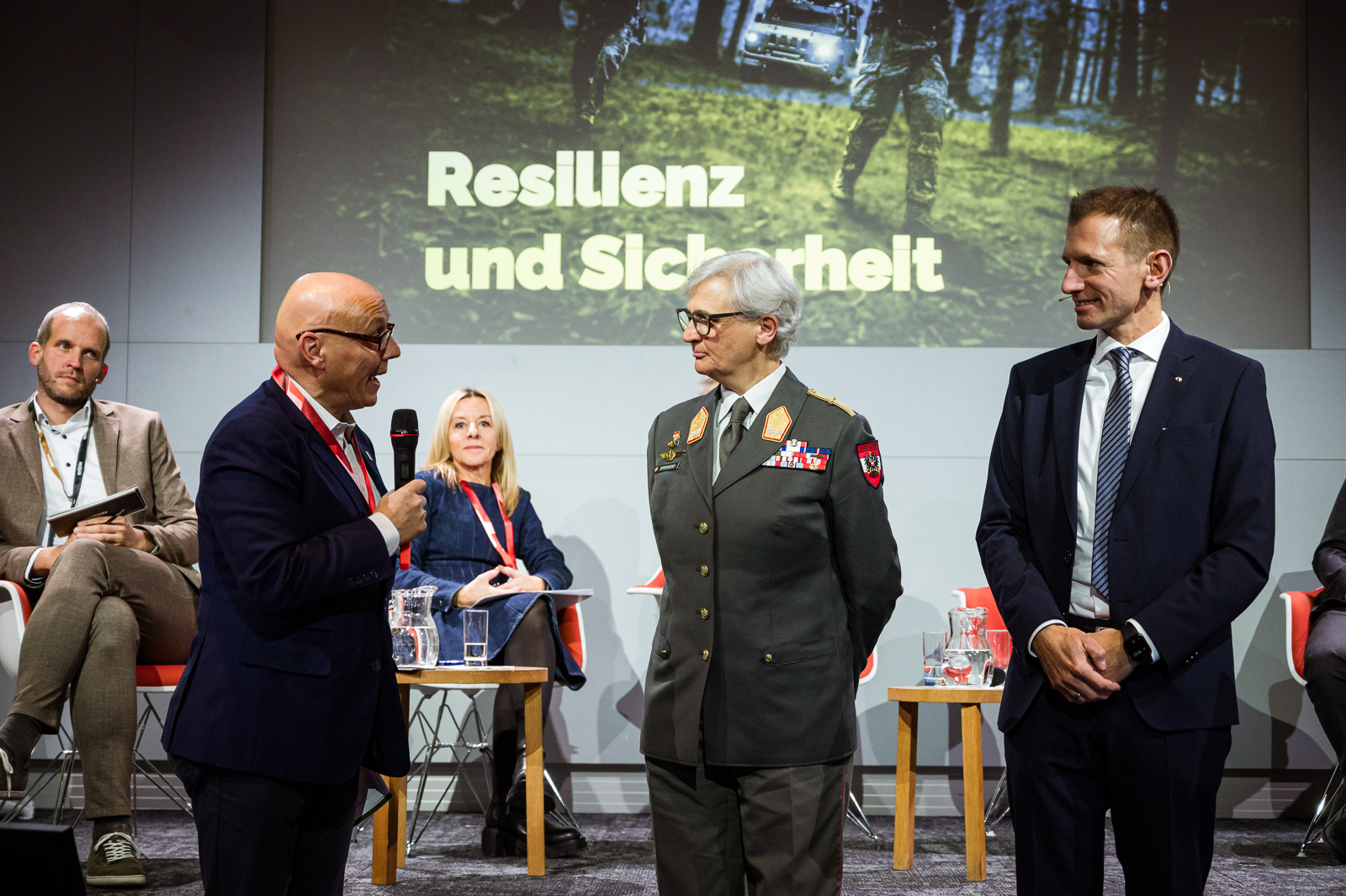 Mann auf Bühne spricht in Handmikrofon. Drei weitere Männer und eine Dame sind ebenfalls auf der Bühne.